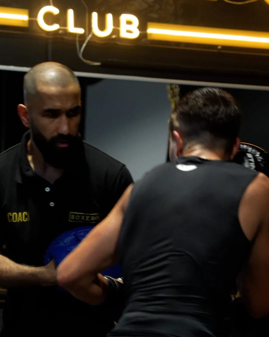 A Boxer’s Club coach holding pads for a member under the neon Club sign
